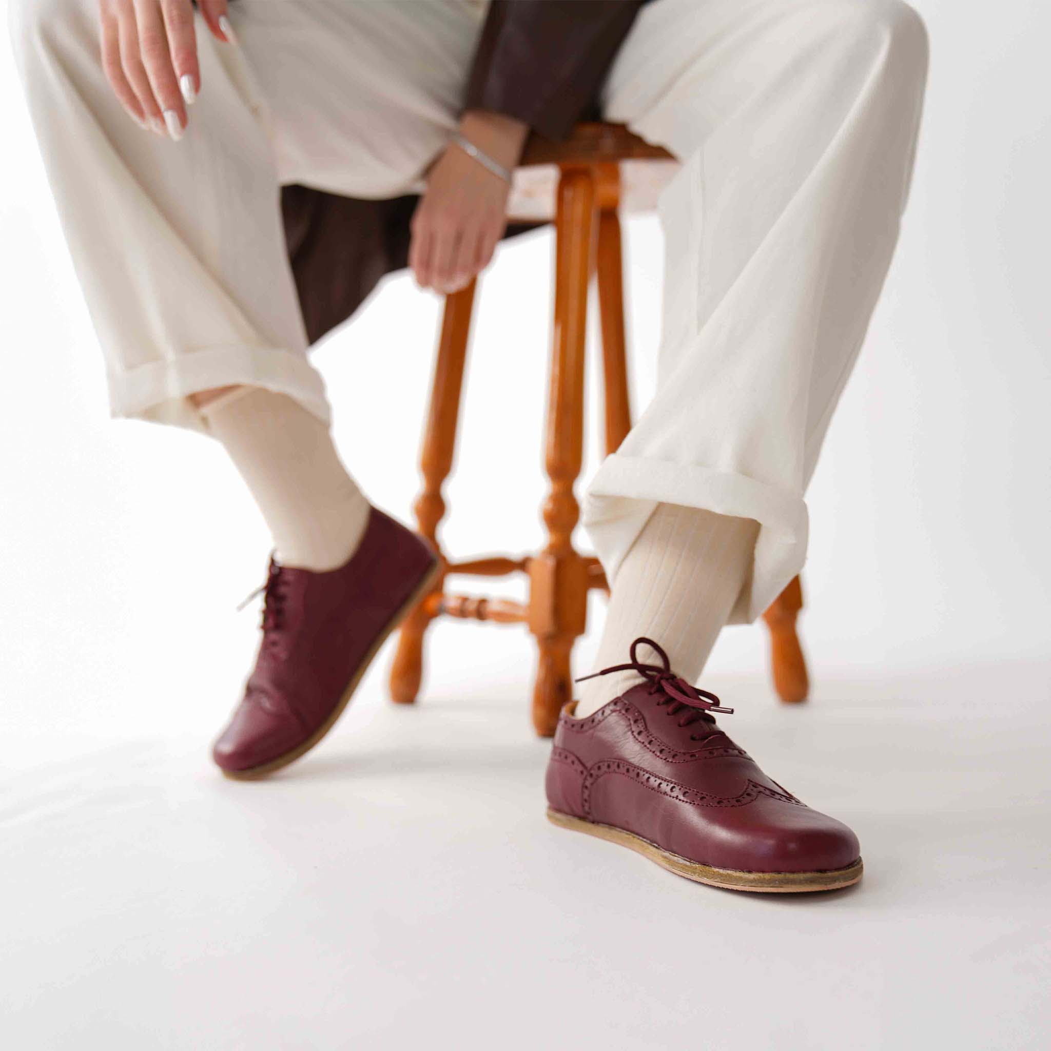Model resting on a wooden stool, highlighting Burgundy Doris Leather Barefoot Women Oxfords.
