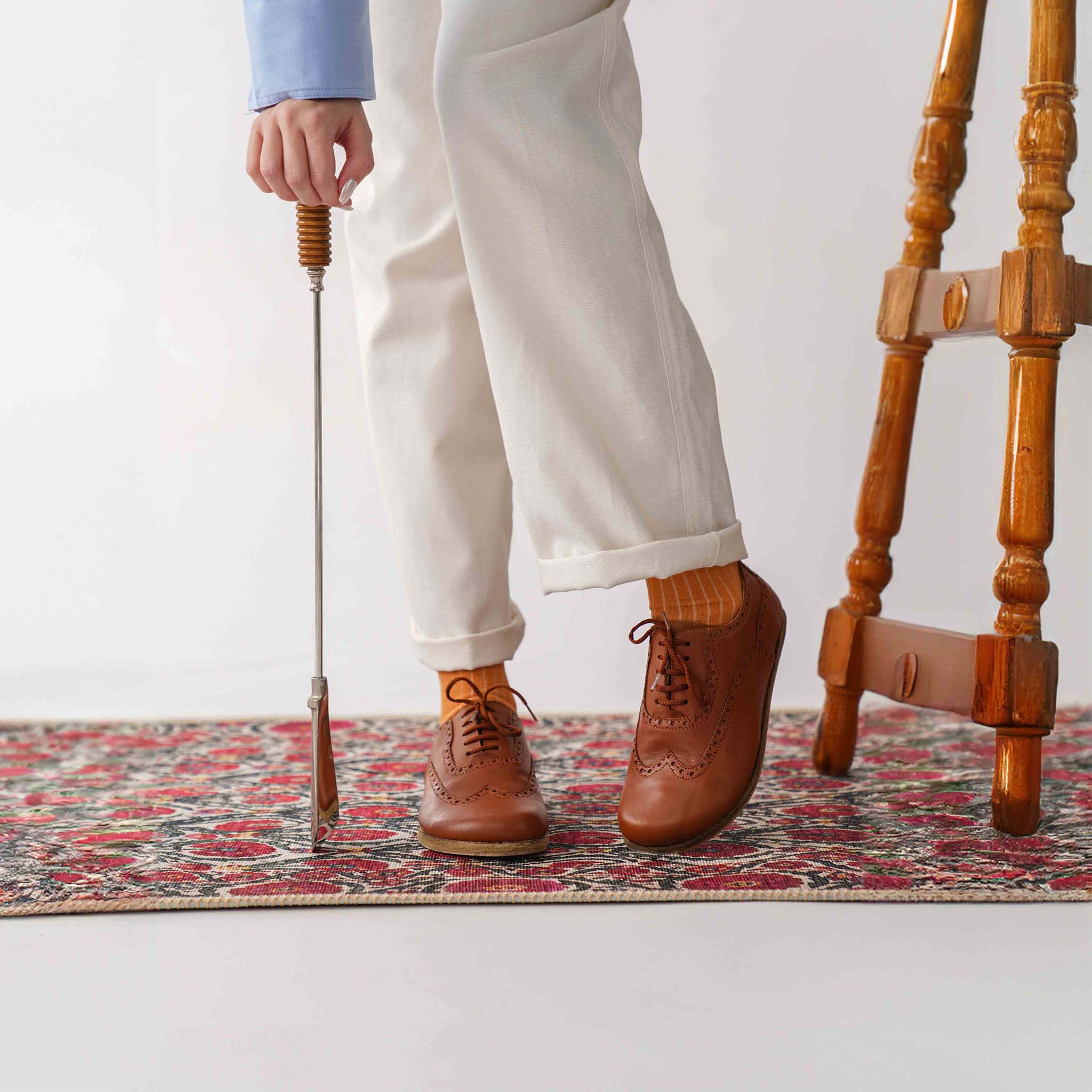 Model adjusting white pants while standing on a patterned rug, wearing tan brown Doris leather barefoot women Oxfords.