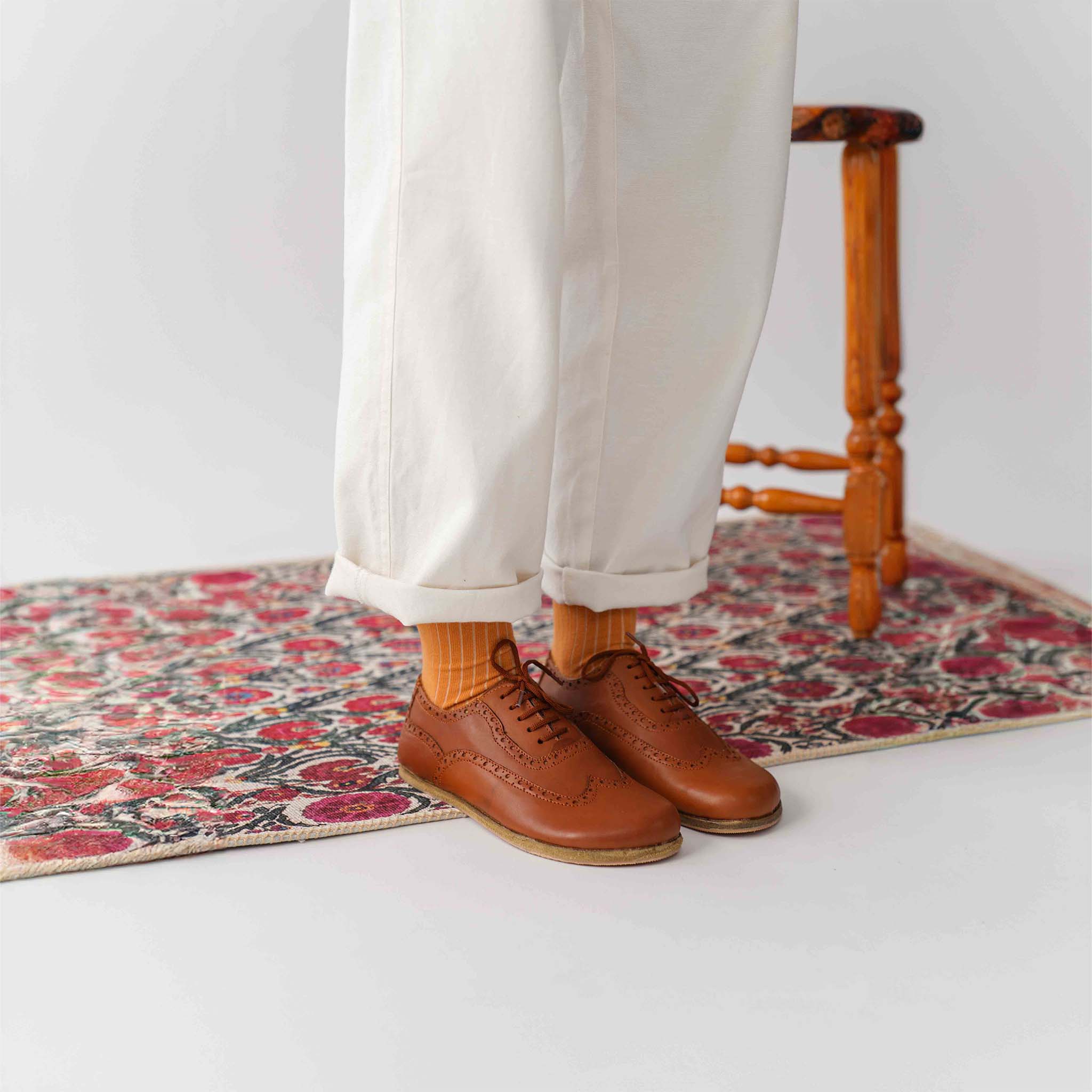 Model sitting on a patterned rug, wearing tan brown Doris leather barefoot women Oxfords with orange socks and white pants.