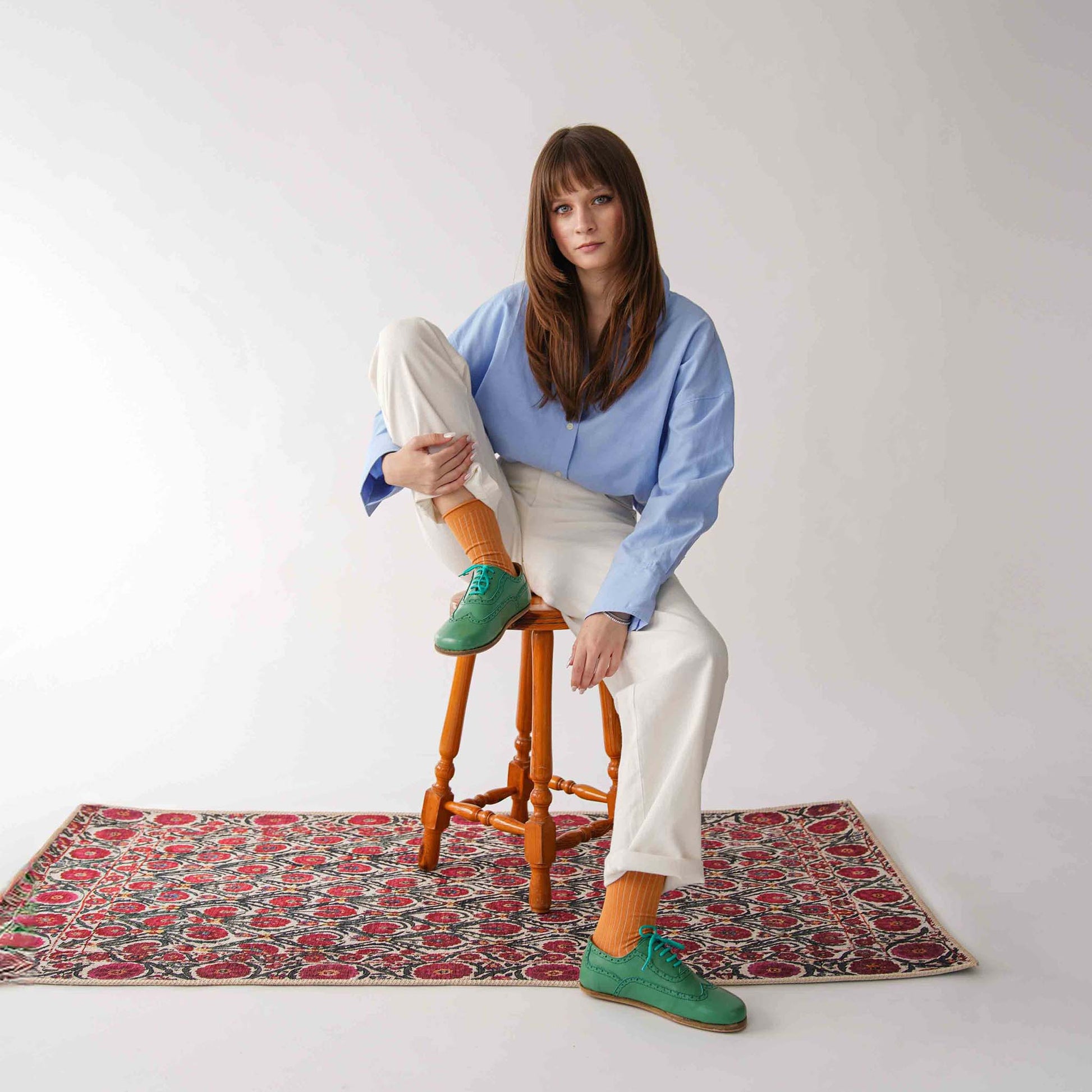 Model sitting on a wooden stool wearing green Doris Leather Barefoot Women Oxfords, paired with white pants and an oversized blue shirt.