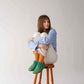 Model sitting on a stool, hugging her knees and wearing green Doris Leather Barefoot Women Oxfords, paired with orange socks and white pants, adding a pop of color to her outfit.