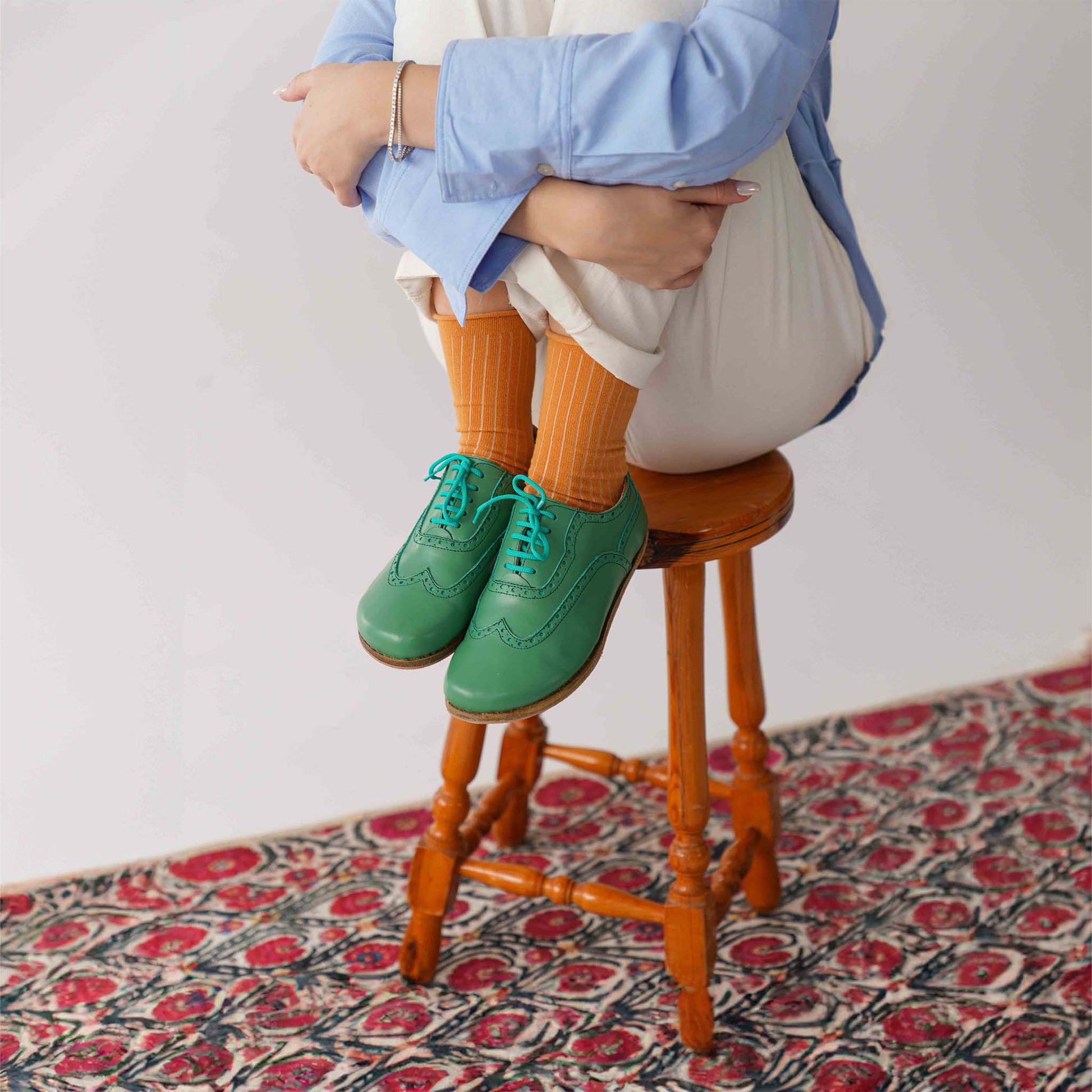 Model wearing green Doris Leather Barefoot Women Oxfords with orange socks, sitting on a stool on a patterned rug.