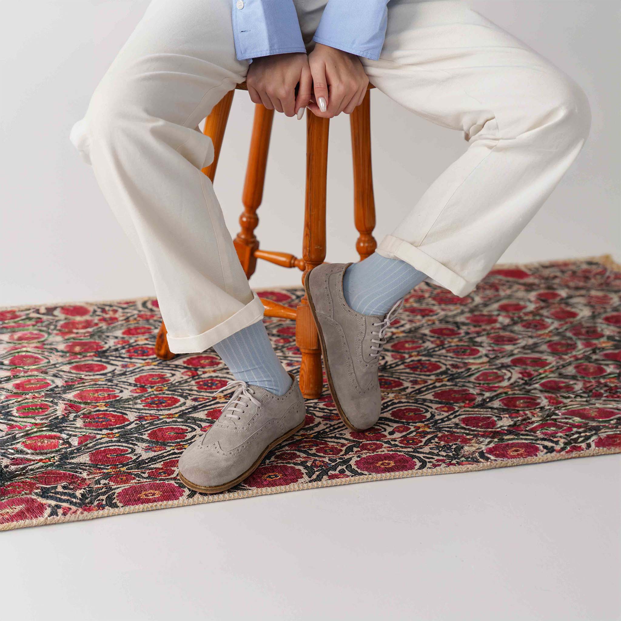 Model wearing gray Doris leather barefoot women Oxfords, styled with white pants and blue socks, seated on a wooden stool.