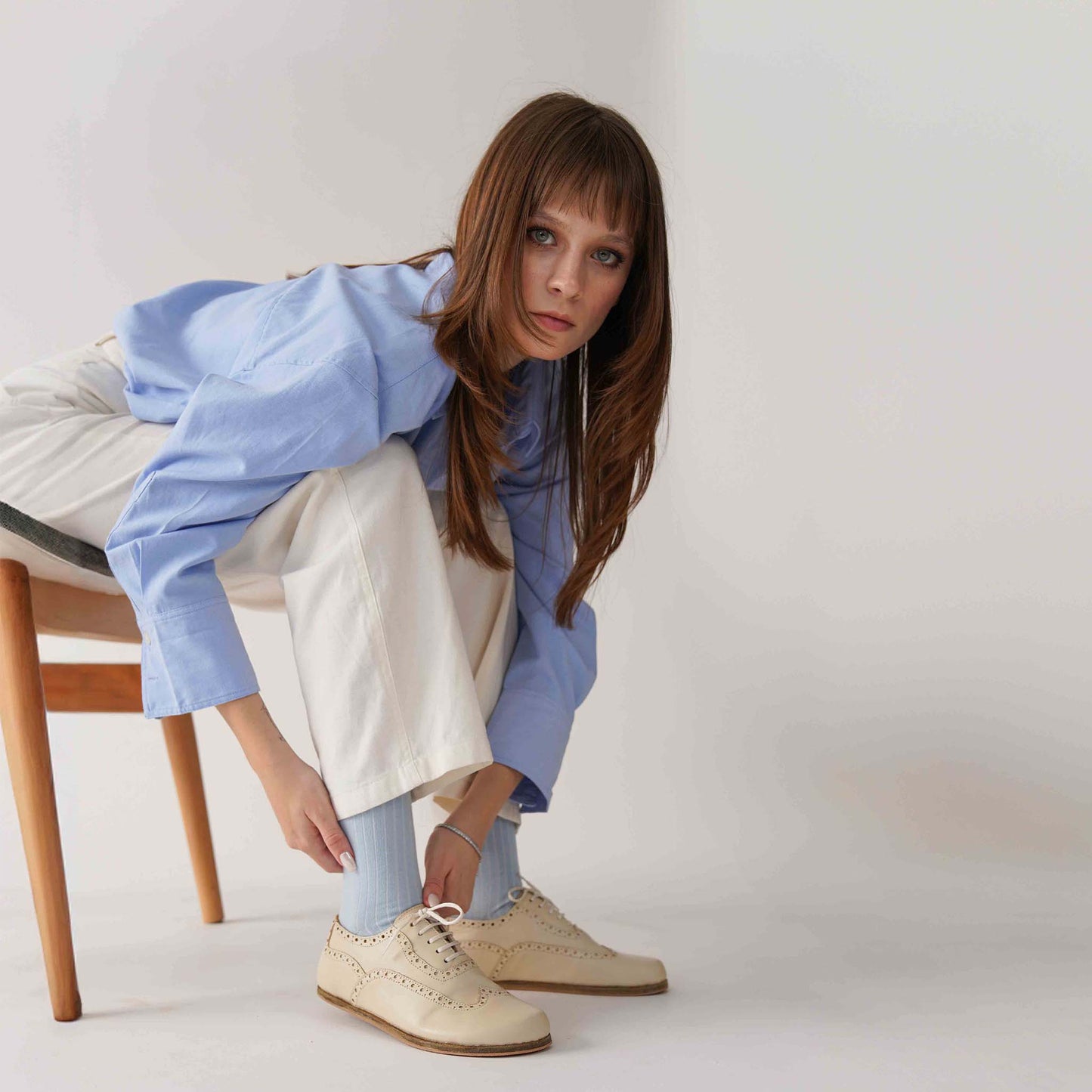 Model adjusting Doris Leather Barefoot Women's Oxfords in beige while sitting on a chair, wearing light blue socks and white pants for a stylish look.