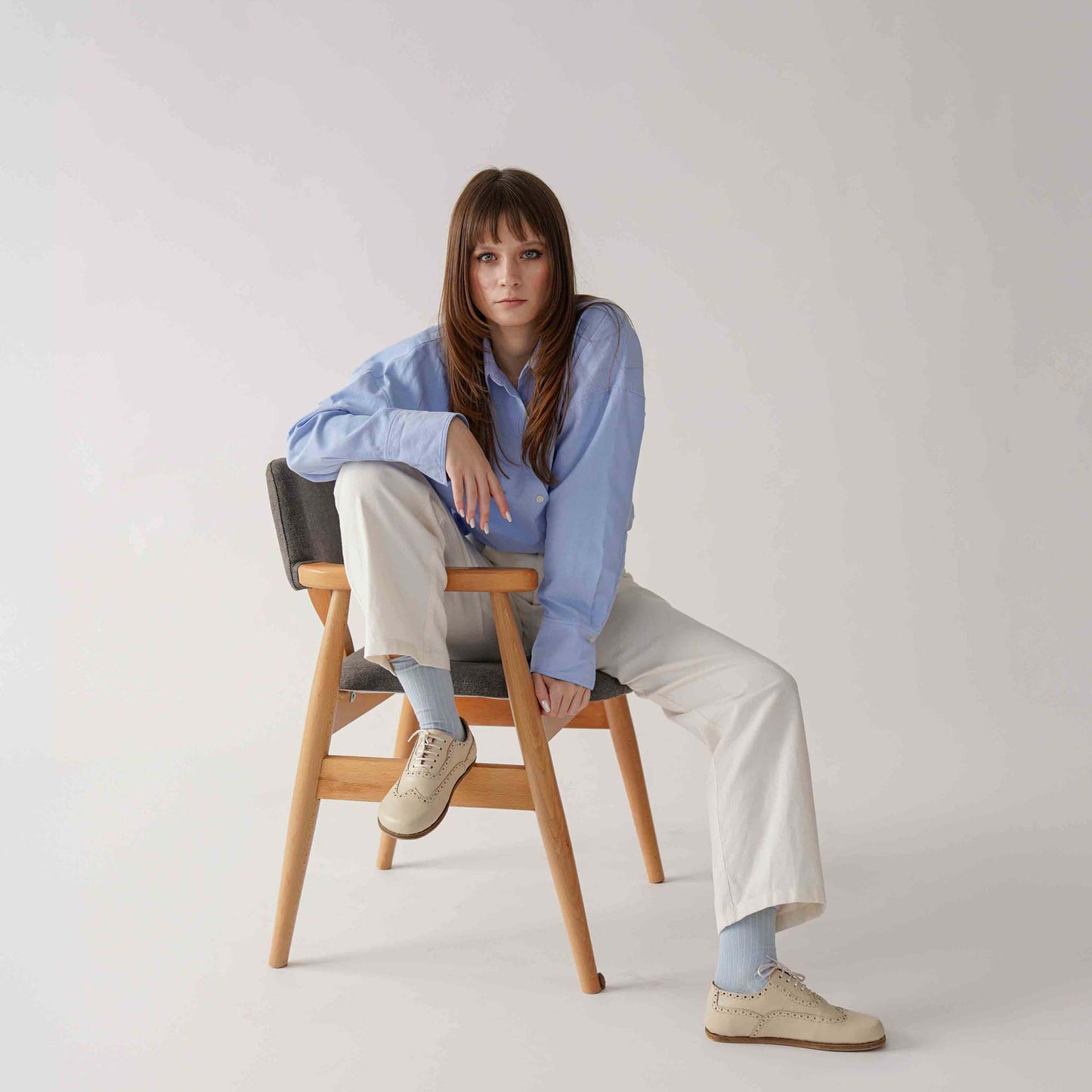 Model sitting on a chair wearing Doris Leather Barefoot Women's Oxfords in beige, paired with light blue socks and white pants.