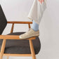 Close-up of a woman resting her foot on a chair while wearing beige Doris leather barefoot women's oxfords with light blue socks.