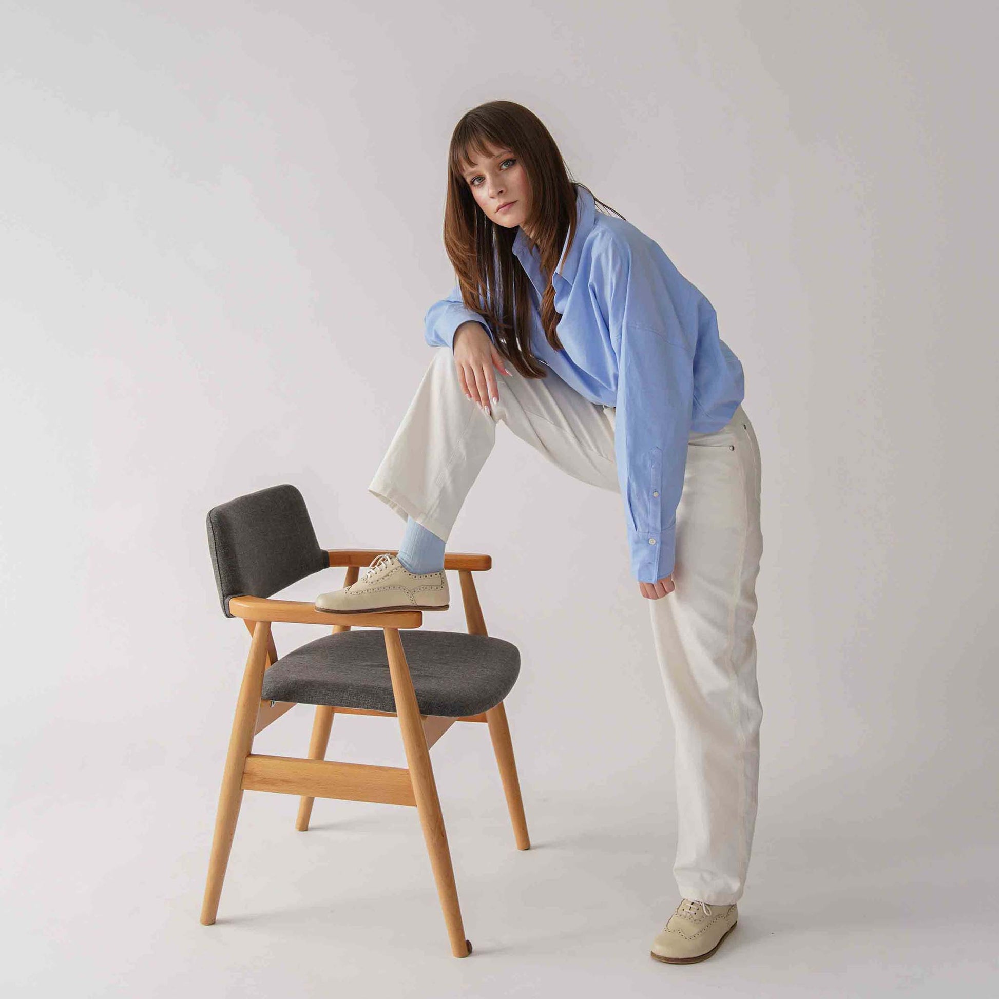 Woman standing with one foot on a chair, wearing beige Doris leather barefoot women's oxfords and a blue shirt with white pants.