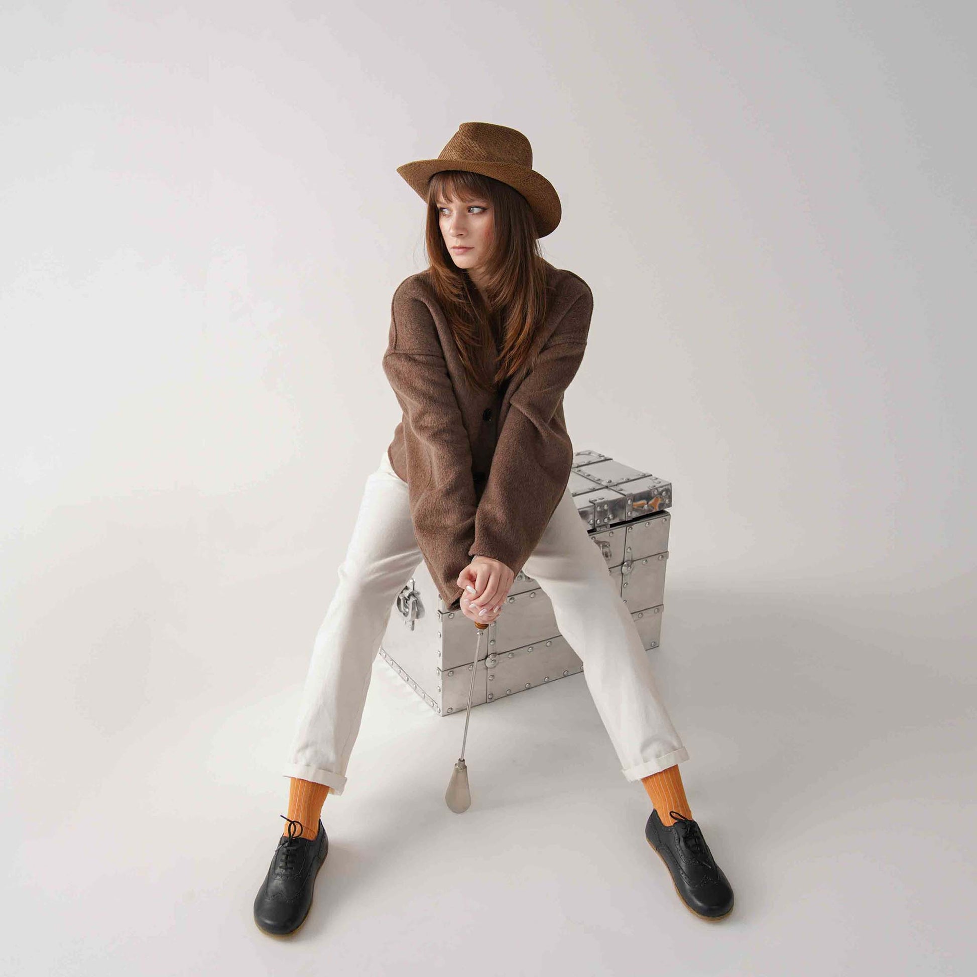 Model wearing black Doris Leather Barefoot Women Oxfords with white pants and a brown coat, sitting on a metallic chest.