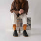 Model wearing black Doris Leather Barefoot Women Oxfords and white pants, seated on a metallic chest with orange socks.