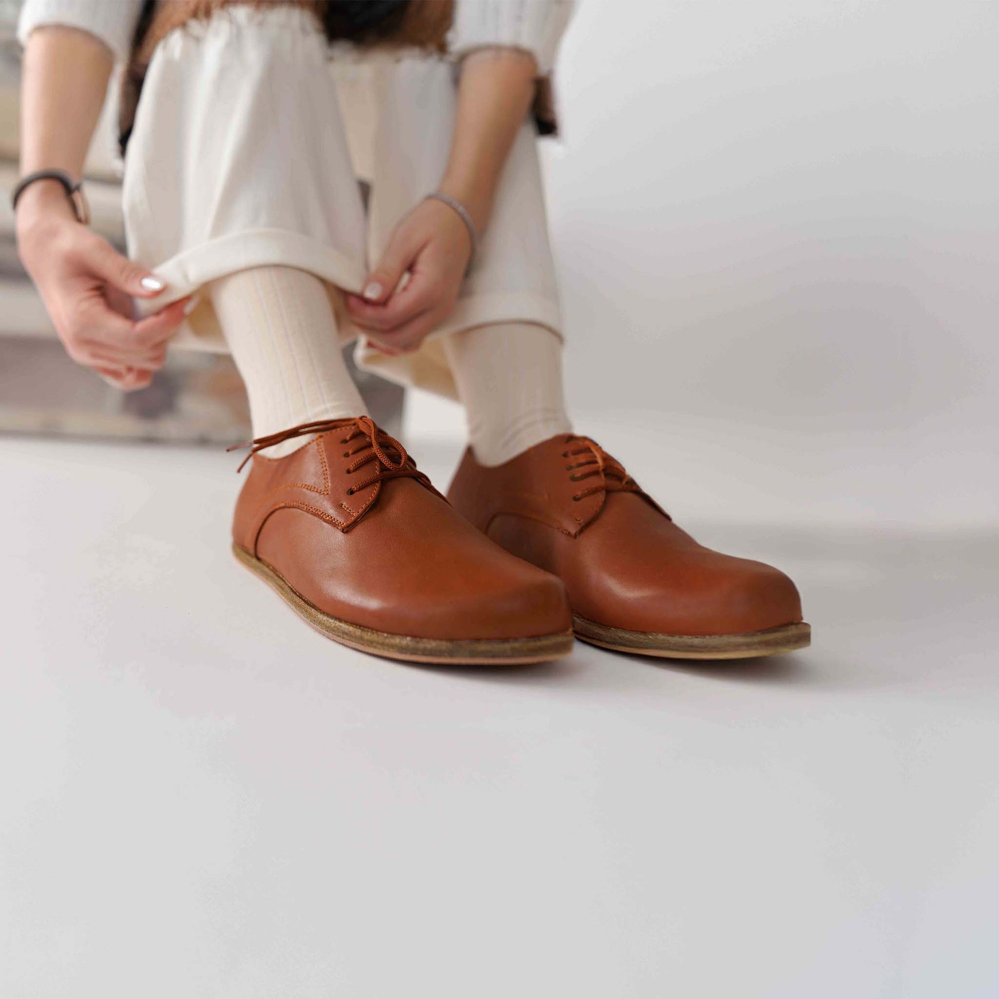 Model adjusting pants, wearing Locris Tan Brown Leather Barefoot Women Oxfords, demonstrating casual and elegant style.