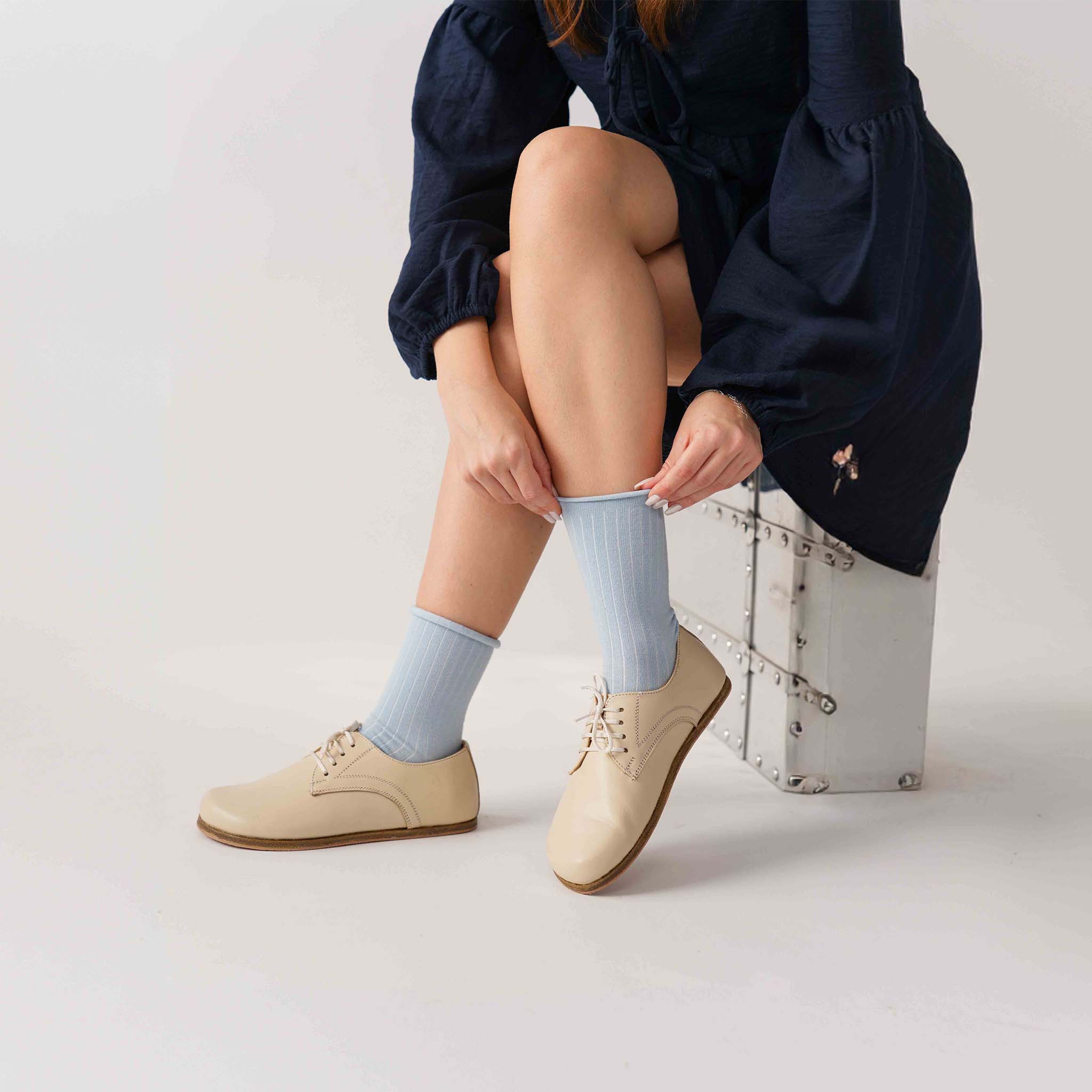 Model adjusting her socks while wearing beige Locris barefoot oxfords, seated on a silver chest.