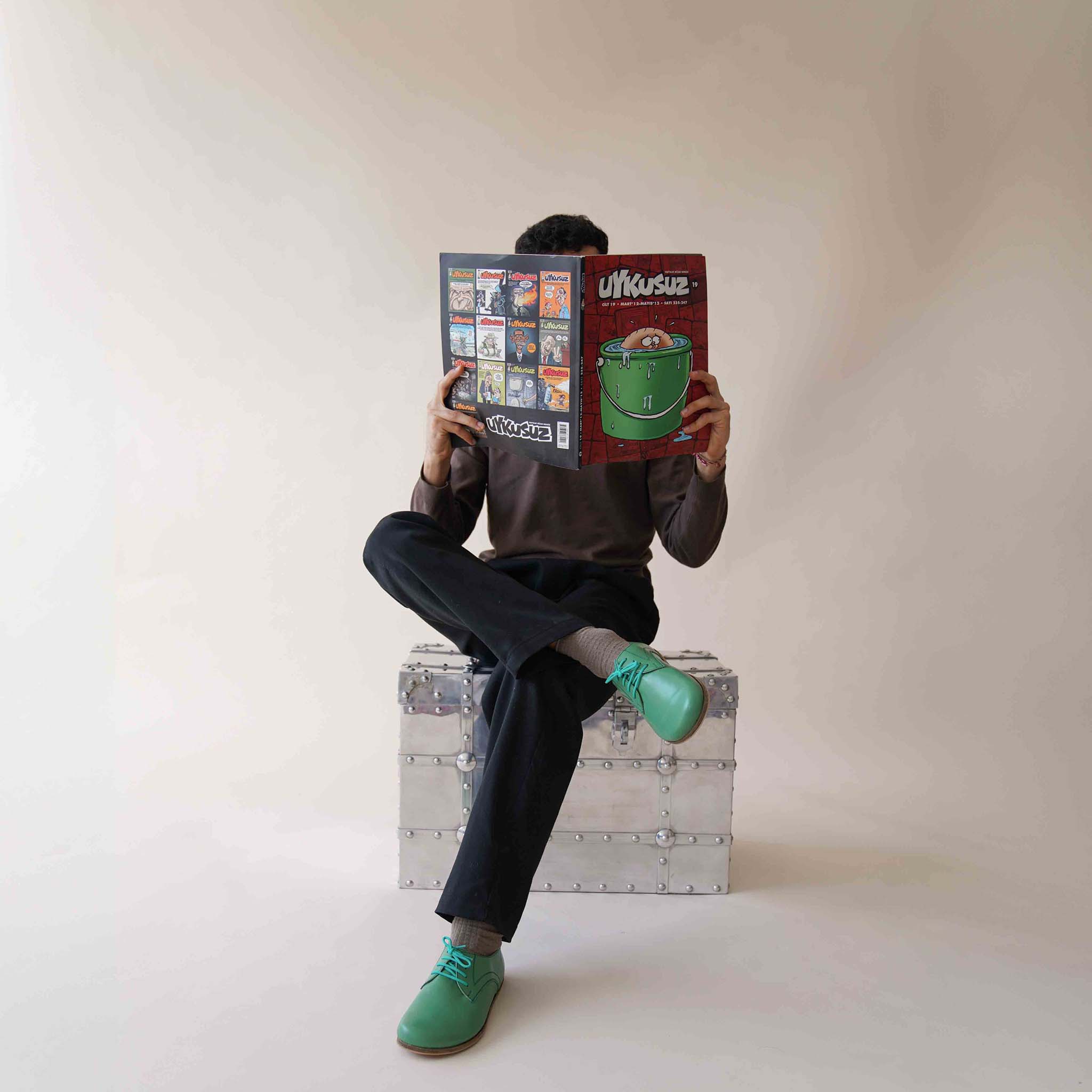 Model sitting with Locris Leather Barefoot Men's Oxfords in green, paired with black pants, showcasing their unique style.