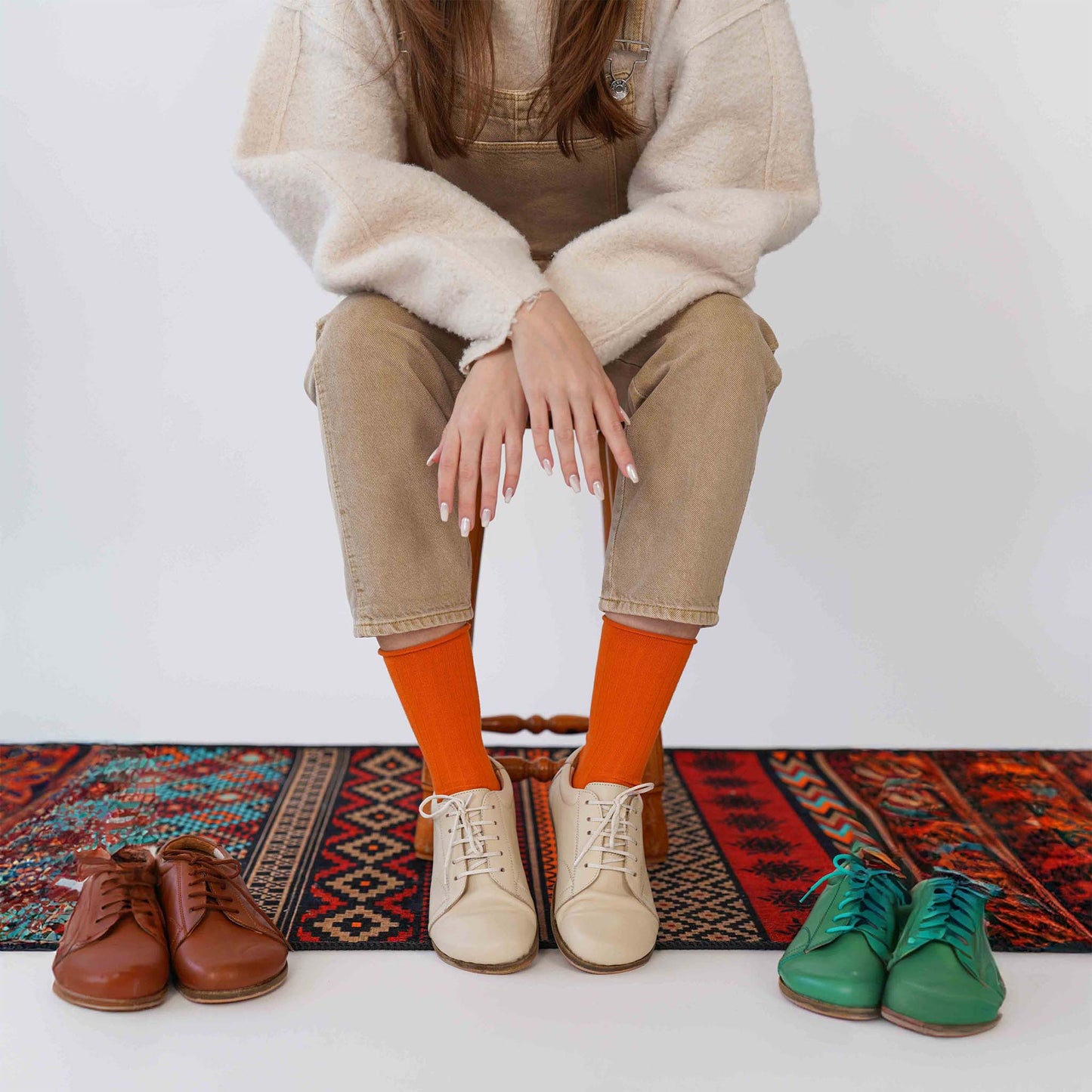 Model sitting with various Lydia leather barefoot women's sneakers in different colors, including green, beige, and brown, on a colorful patterned rug.