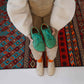 Model holding green Lydia leather barefoot women's sneakers with turquoise laces, sitting on a patterned rug, emphasizing the shoes' vibrant color and natural fit.