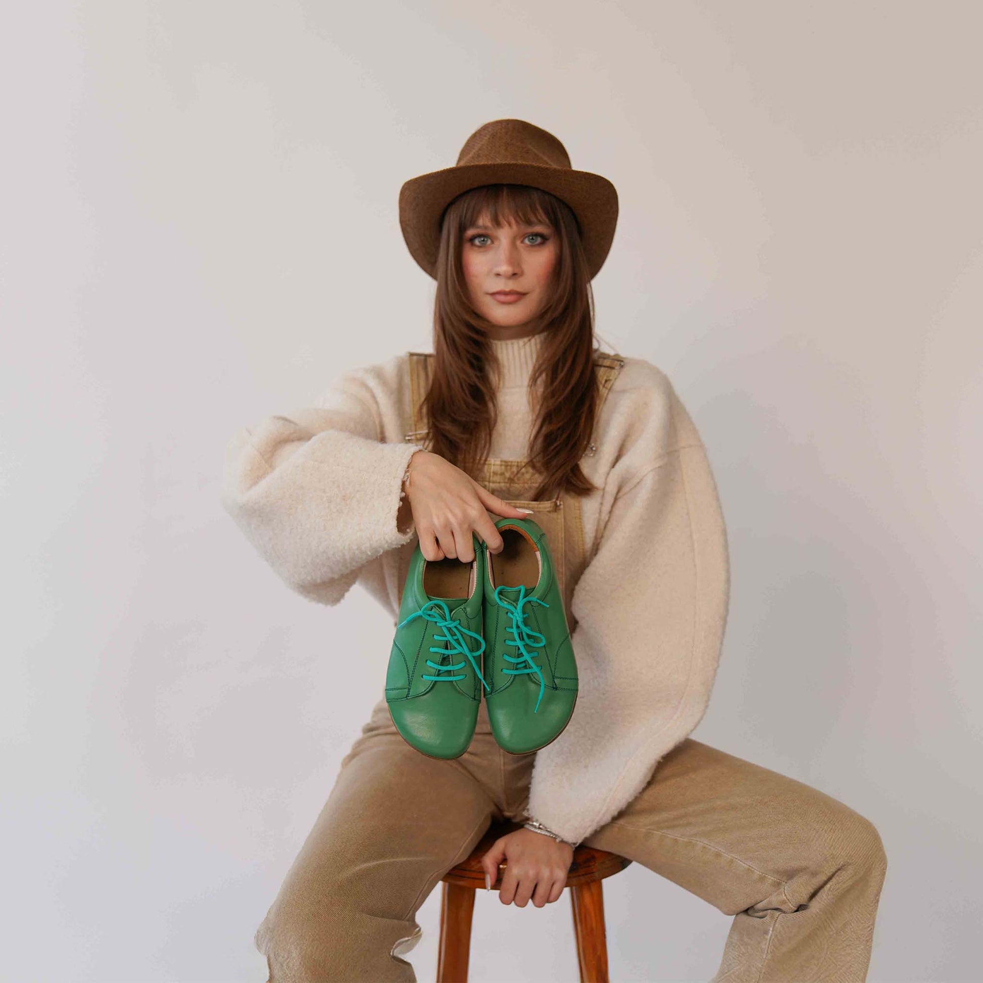 Model holding a pair of Lydia Leather Barefoot Women Sneakers in green with turquoise laces, perfect for casual outfits.