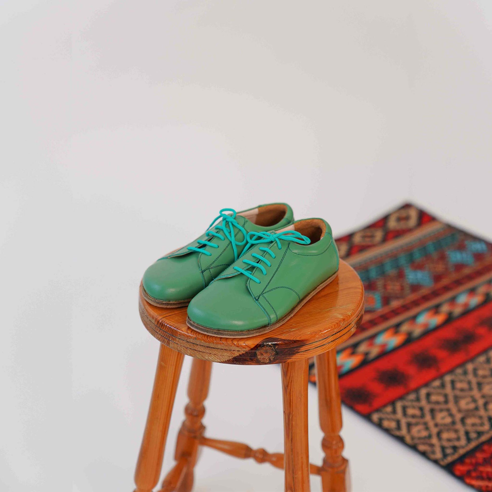 Green Lydia leather barefoot women's sneakers with turquoise laces, placed on a wooden stool, highlighting the shoes' stylish design and high-quality leather.