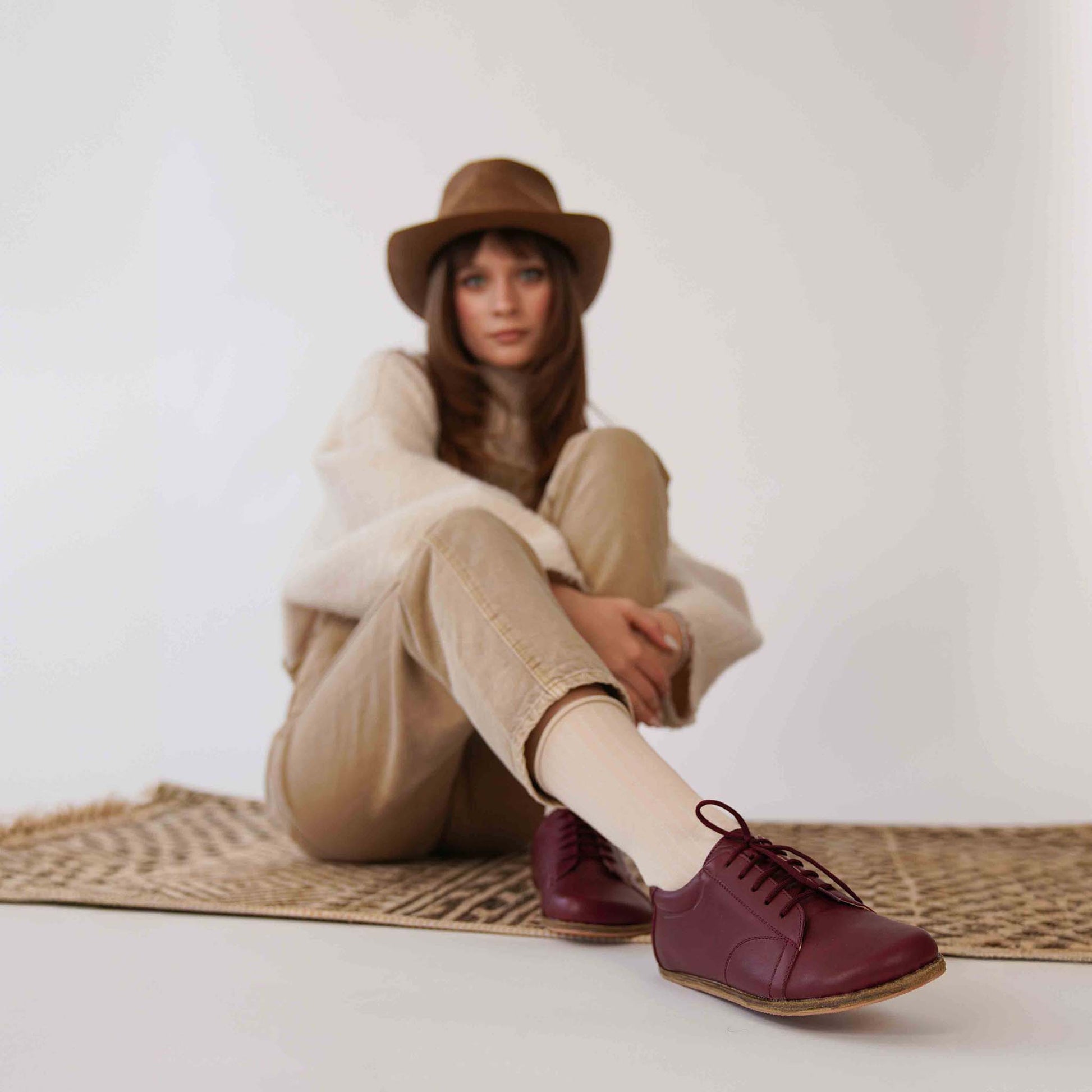 Woman sitting on a patterned rug wearing burgundy Lydia leather barefoot sneakers, paired with beige pants and a cozy sweater.