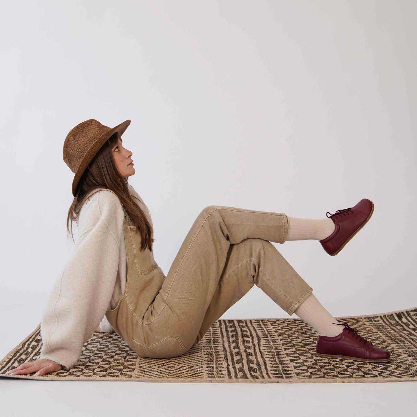 Woman lounging in beige overalls and a sweater, wearing burgundy Lydia leather barefoot sneakers, emphasizing casual and comfortable style.