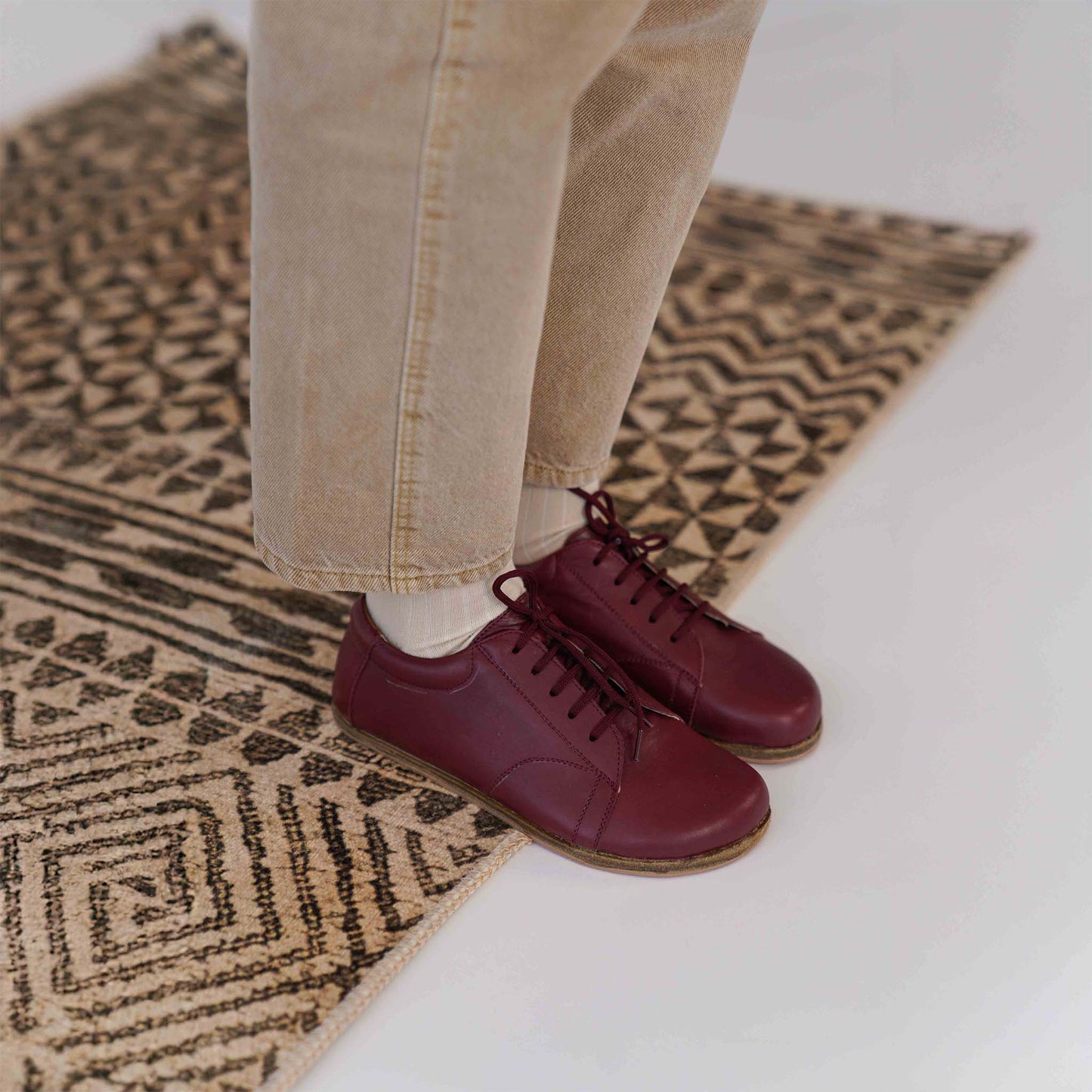 Burgundy barefoot sneakers showcased on a patterned rug, demonstrating stylish design and natural foot alignment.