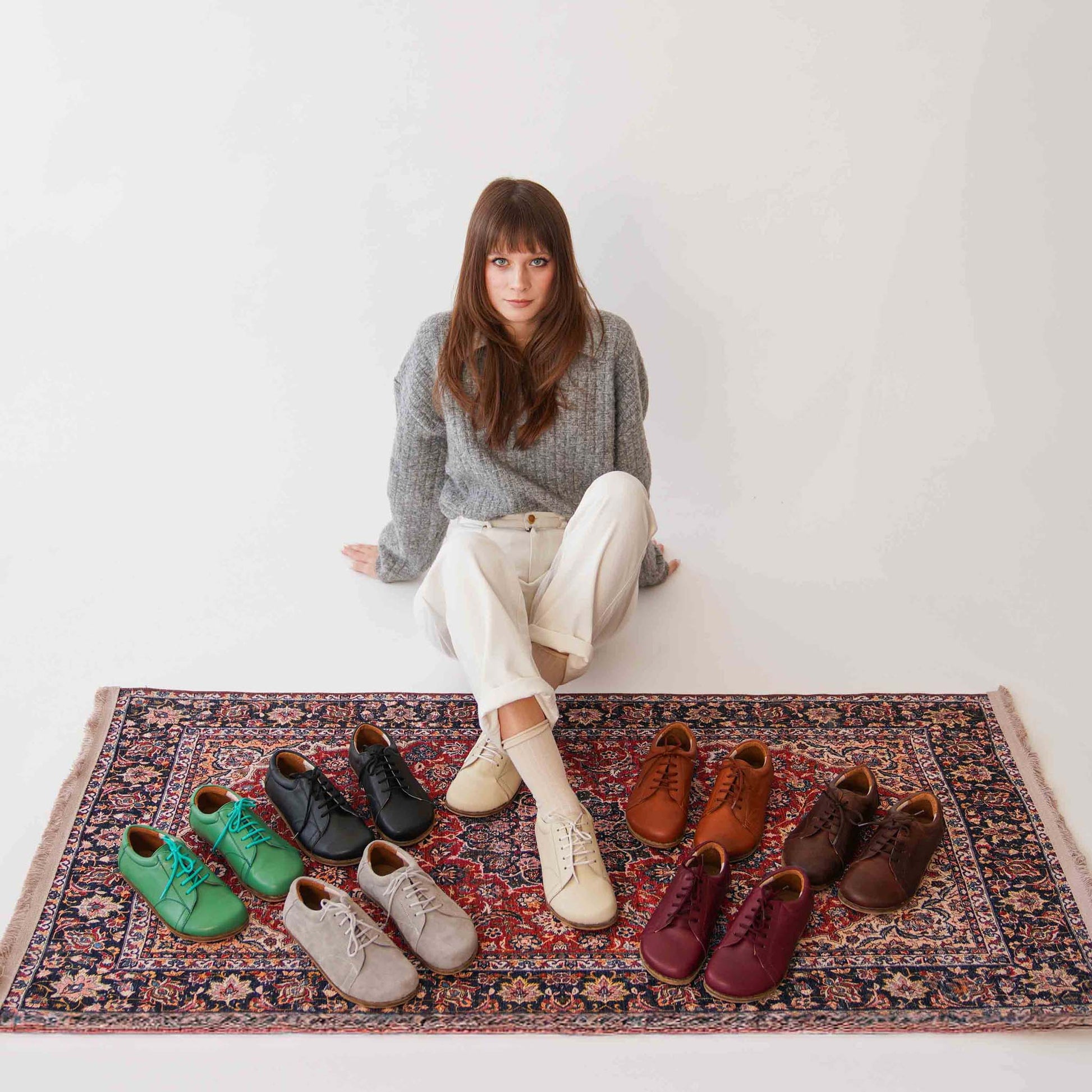 Model seated on a vibrant rug, wearing beige leather barefoot women's sneakers, portraying a blend of fashion and comfort.