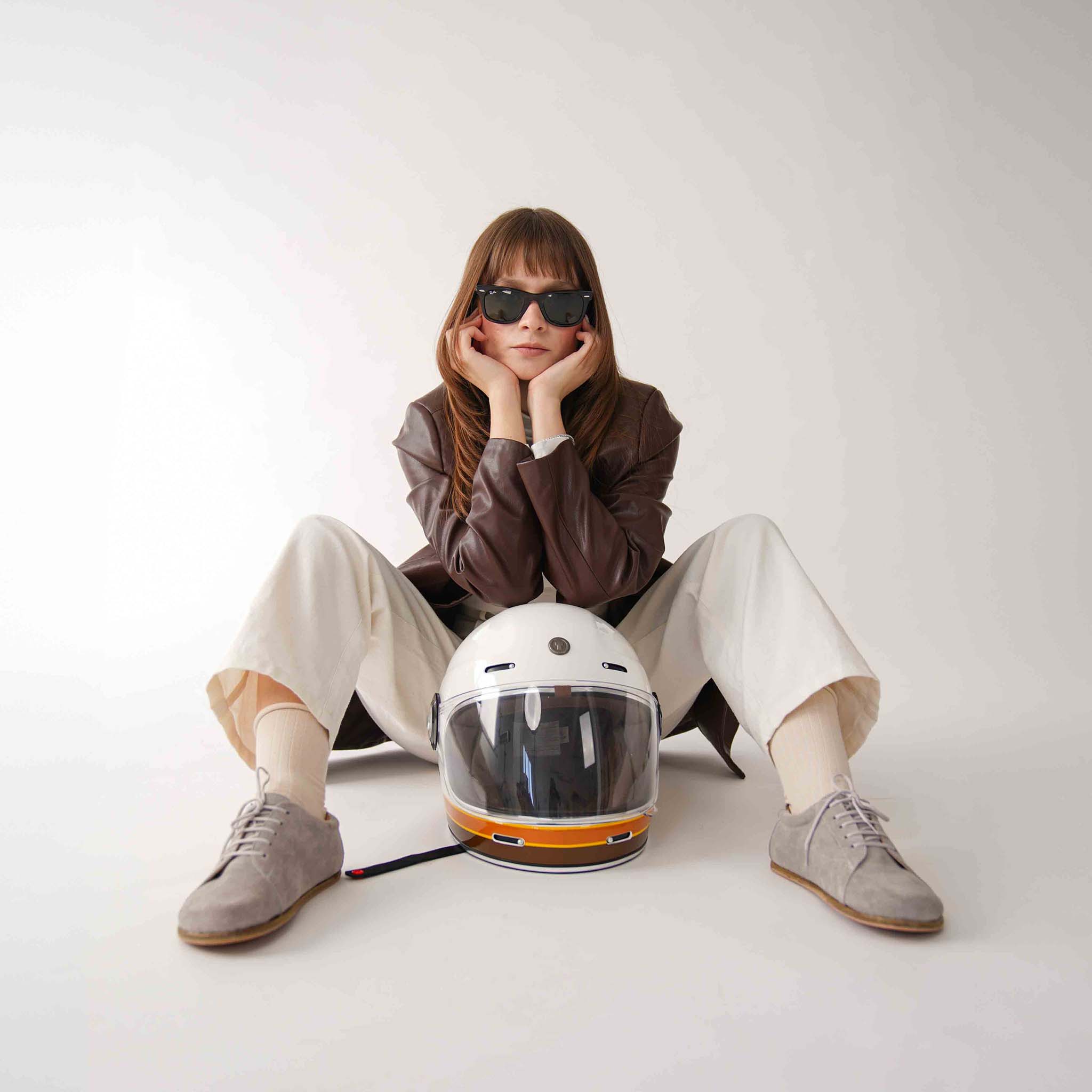 Model wearing gray suede barefoot sneakers, paired with a leather jacket and helmet, showcasing a casual and stylish look.