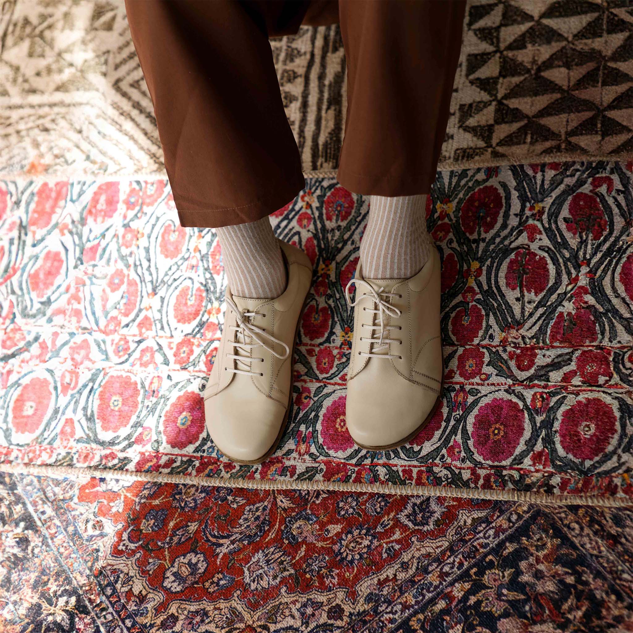 Lydia Leather Barefoot Men's Sneakers in beige, modeled on a colorful rug, highlighting their versatile and stylish look.