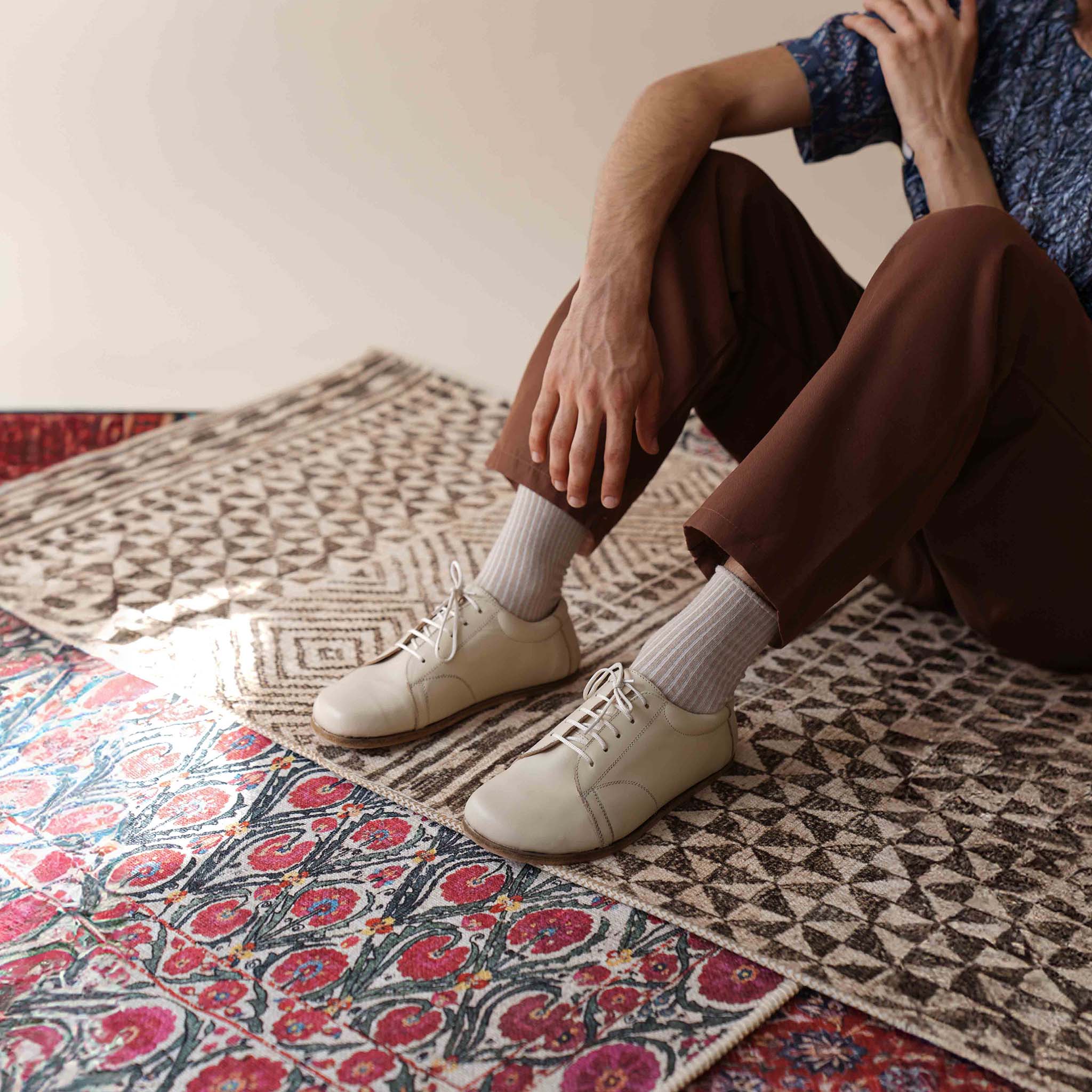 Model sitting on a patterned rug, wearing Lydia Leather Barefoot Men's Sneakers in Beige, ideal for casual comfort.