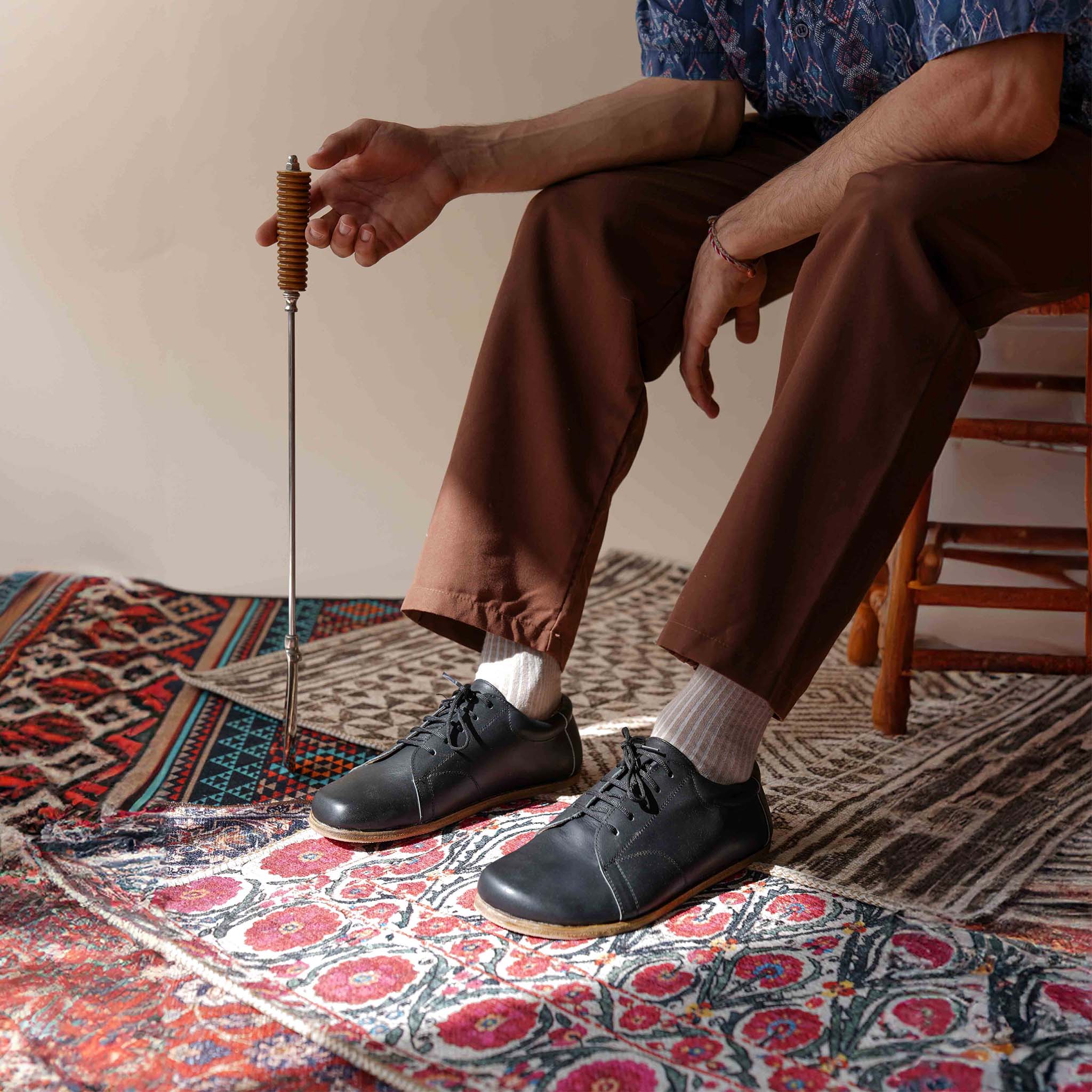 Model sitting on a patterned rug, showcasing the Black Lydia Leather Barefoot Men's Sneakers with a classic lace-up design.