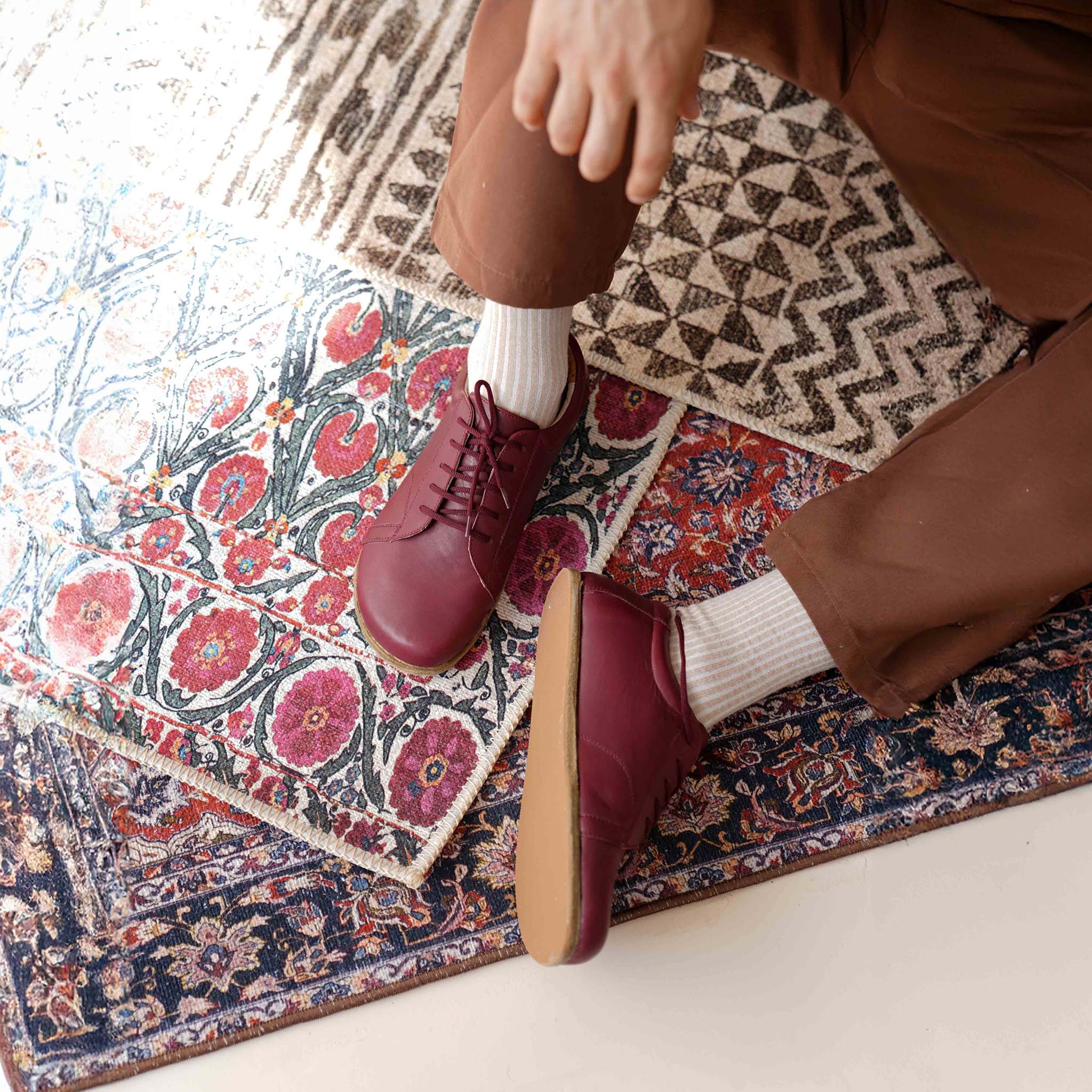 Burgundy Lydia Leather Barefoot Men's Sneakers displayed on a vibrant patterned rug, highlighting the contrast and stylish look.